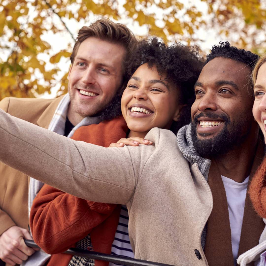 Group Of Friends Outdoors Wearing Coats And Scarves Posing For Selfie On Phone In Autumn Park
